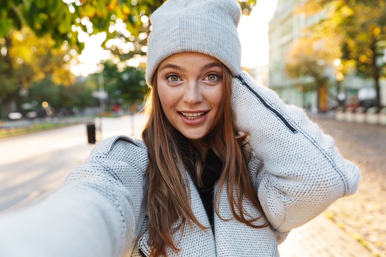 Beautiful young woman dressed in autumn coat and hat walking outdoors, taking a selfie