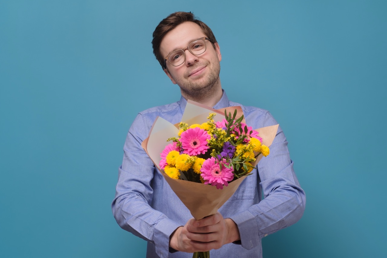 Young caucasian man in pink t-shirt holding flowers as gift for his mother or girlfriend on birthday. Anniversary present concept. Studio shot on colored wall.